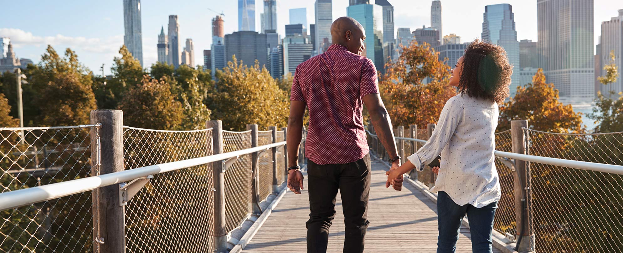 A man and woman holding hands walk along a bridge with fall-colored trees and a city skyline in the distance. 